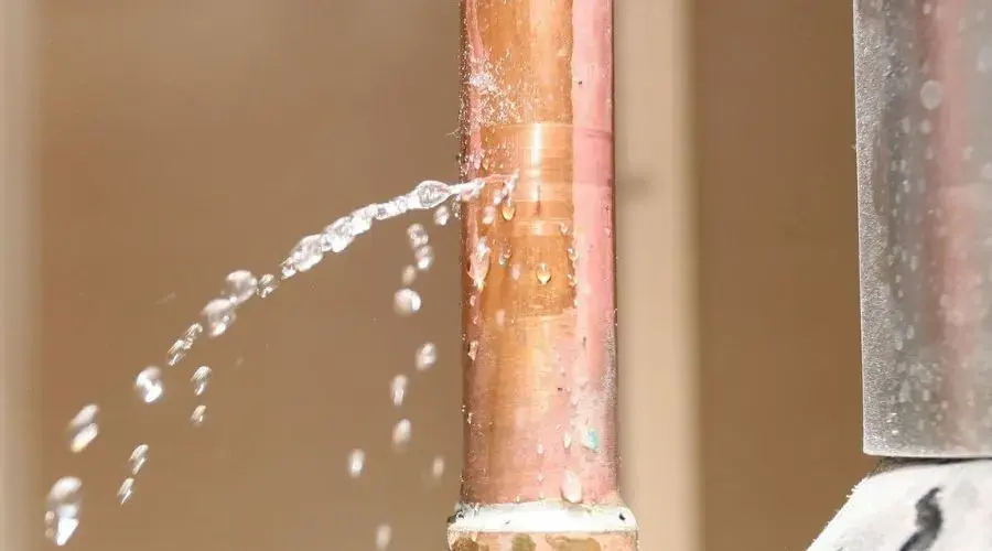 Water spraying from a split in a copper pipe along a wall, burst pipe damage in Anoka